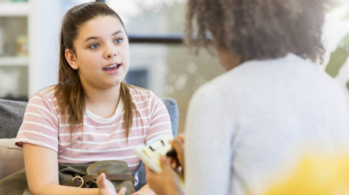 A serious teenage girl gestures as she sits on a couch in her school counselor's office and talks to her unrecognizable counselor.  The counselor takes notes on a clipboard.