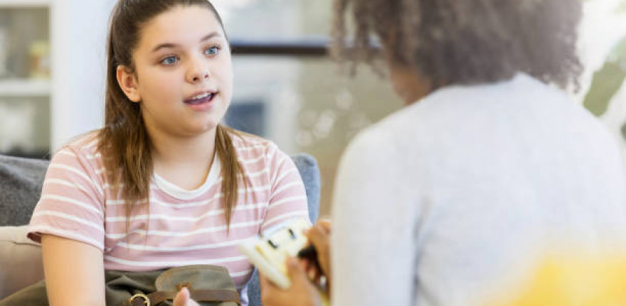 A serious teenage girl gestures as she sits on a couch in her school counselor's office and talks to her unrecognizable counselor.  The counselor takes notes on a clipboard.