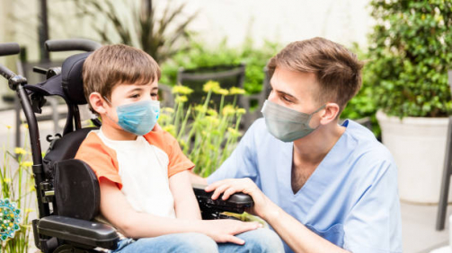 Portrait of beautiful little boy on wheelchair and friendly nurse at the hospital both wearing protective facemasks - Pandemic lifestyles