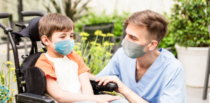 Portrait of beautiful little boy on wheelchair and friendly nurse at the hospital both wearing protective facemasks - Pandemic lifestyles